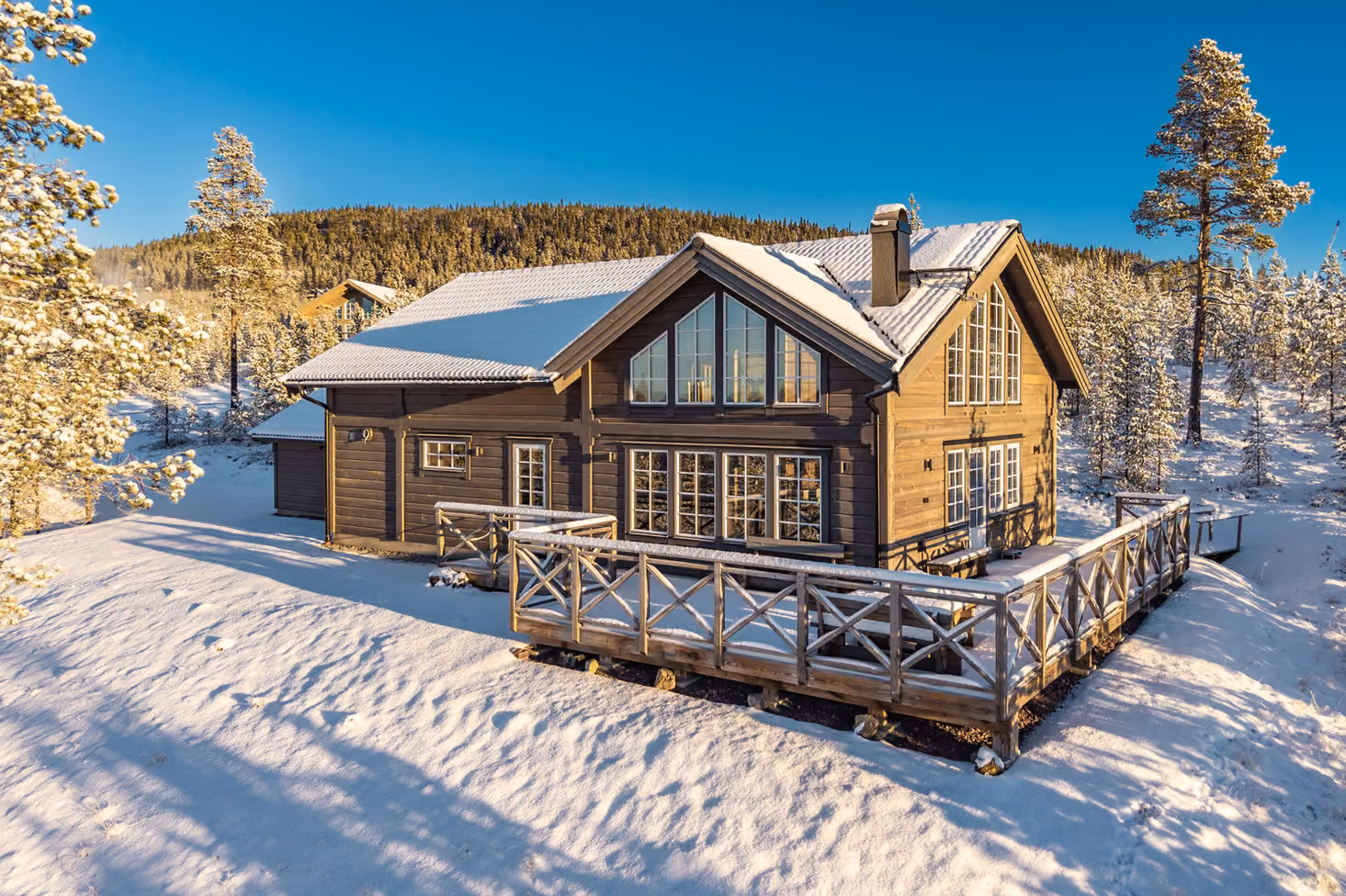 Wooden cabin covered in snow with a deck, surrounded by snow-covered trees under a clear blue sky.