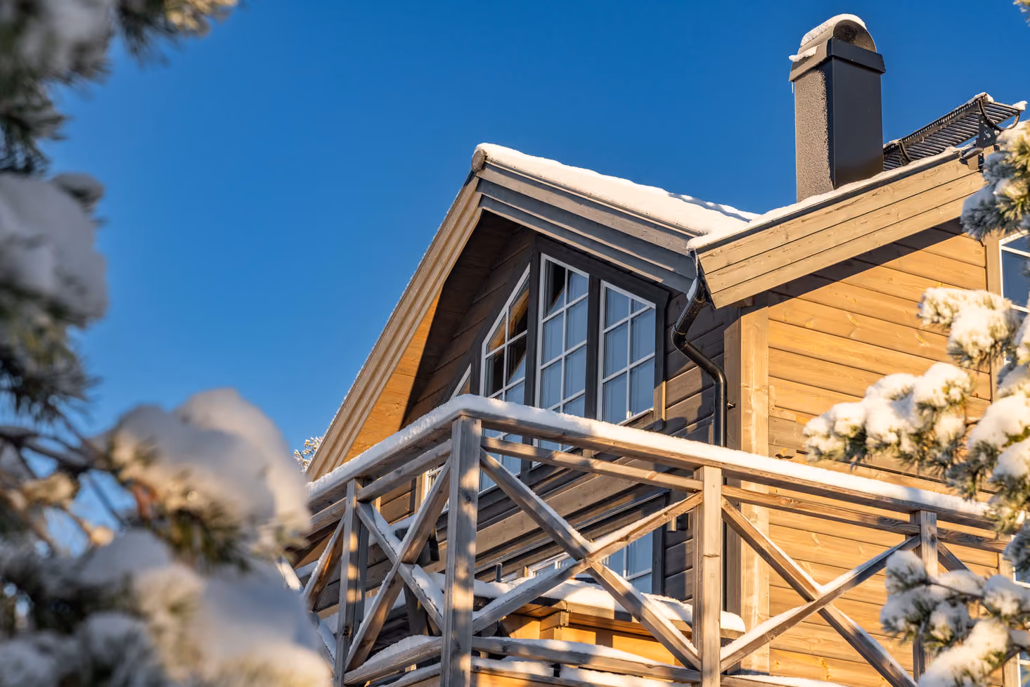 Snow-covered wooden balcony and roof of a house with blue sky and snow-covered trees around.
