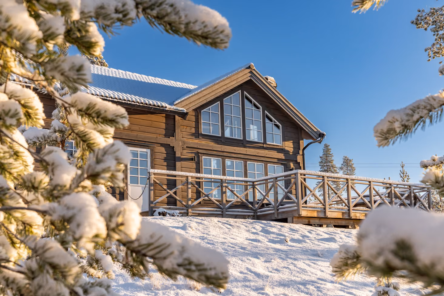 Wooden cabin with snow-covered roof and deck surrounded by snow and pine trees under blue sky.