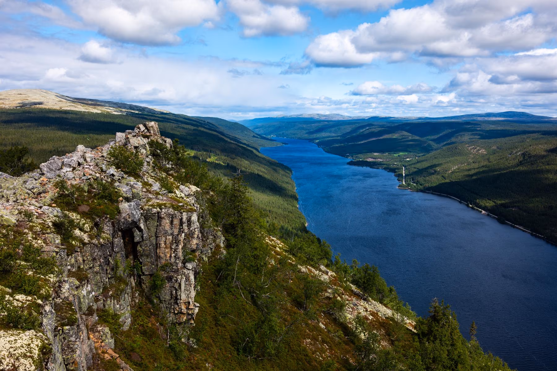 Wide river flowing through forested mountains with rocky cliffs under a partly cloudy sky.