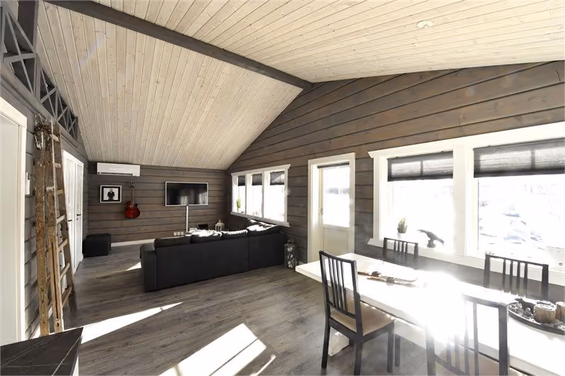 Bright modern living room with wood-paneled walls, black sofa, wall TV, and dining table by large windows.