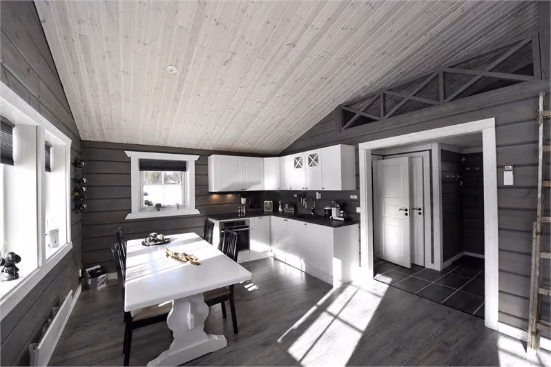 Modern kitchen with white cabinets, dining table, dark walls, wood ceiling, and sunlight through windows.