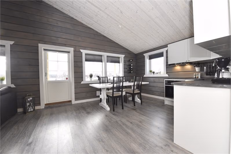 Modern dining area with white table, four black chairs, dark wood walls, and white kitchen cabinets.