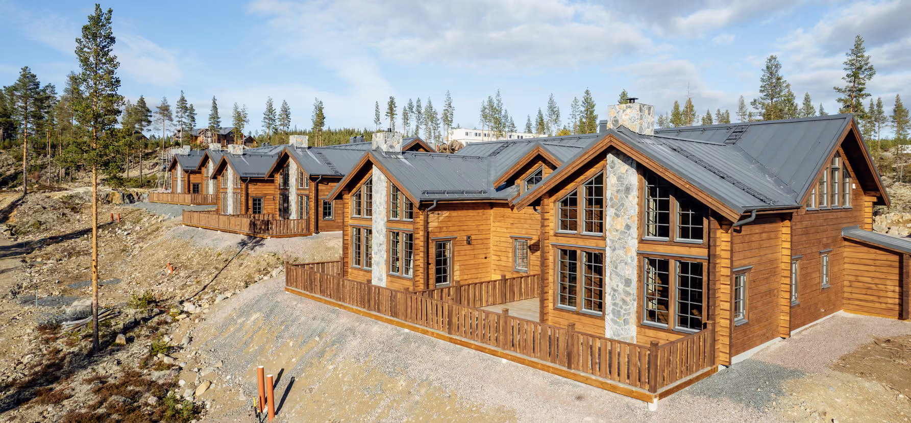 Row of modern wooden cabins with stone chimneys and fenced patios in a forested, rocky area.