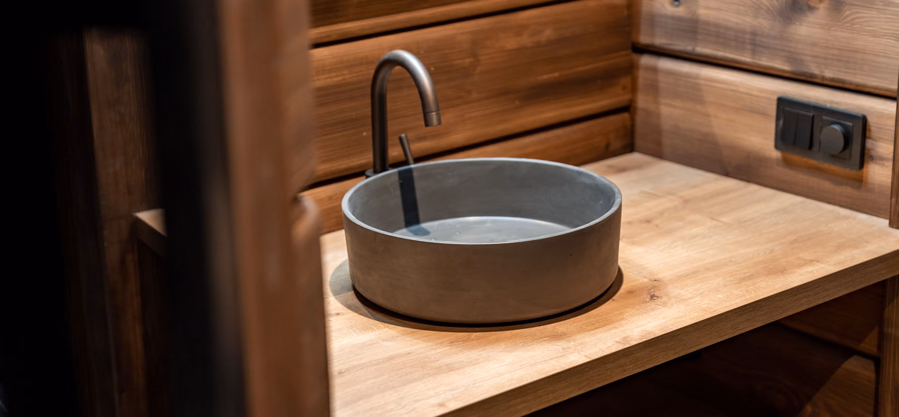 Round concrete sink with a brushed metal faucet on a wooden countertop in a wood-paneled room.