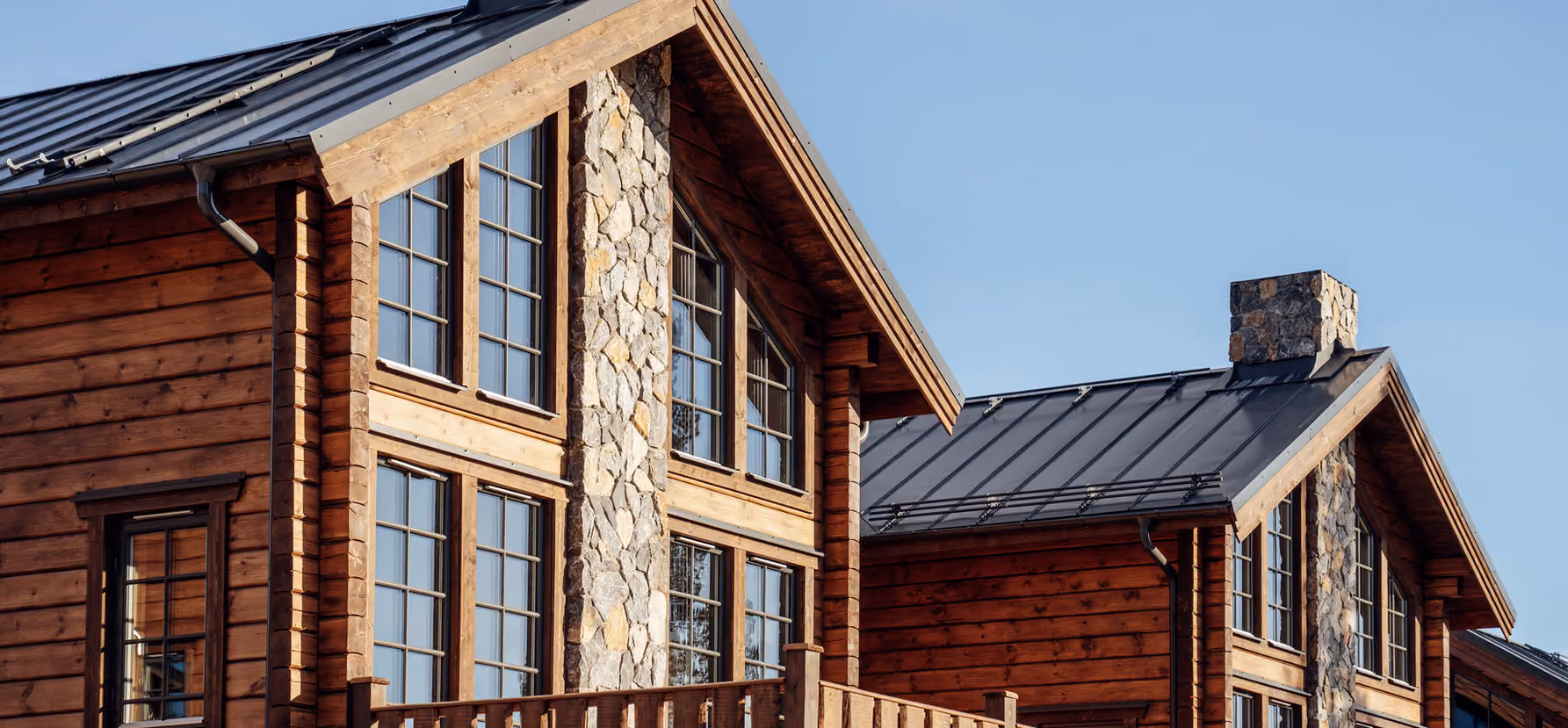 Exterior of rustic-style wooden house with large windows, stone accents, and metal roof under clear sky.