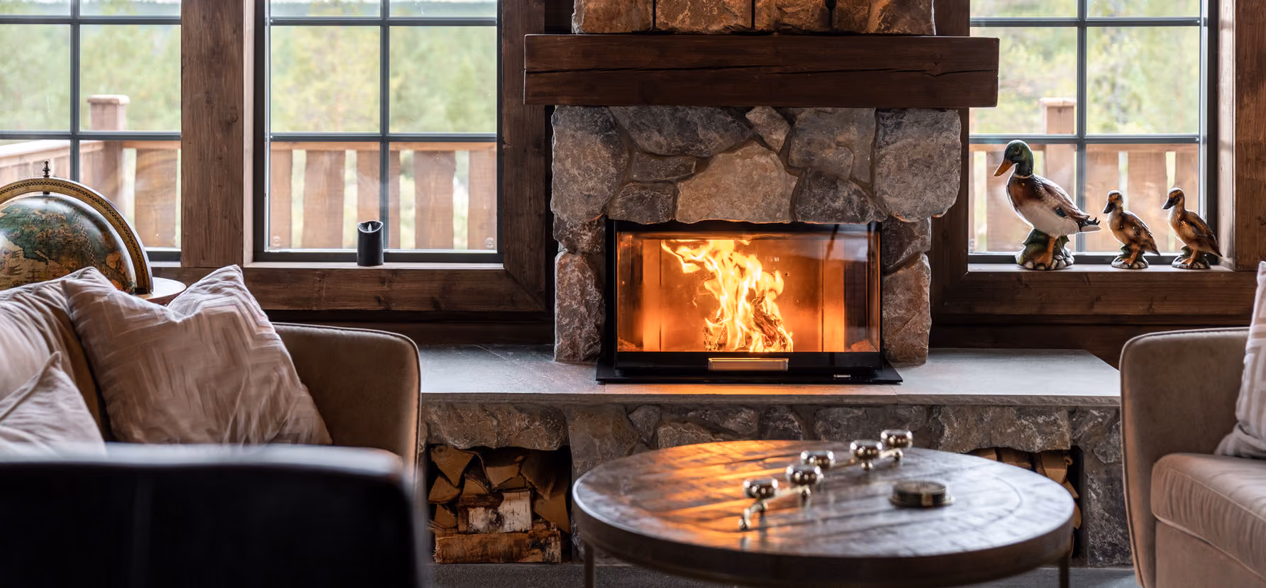 Cozy living room with stone fireplace, burning fire, wooden table, globe, and duck figurines on windows.