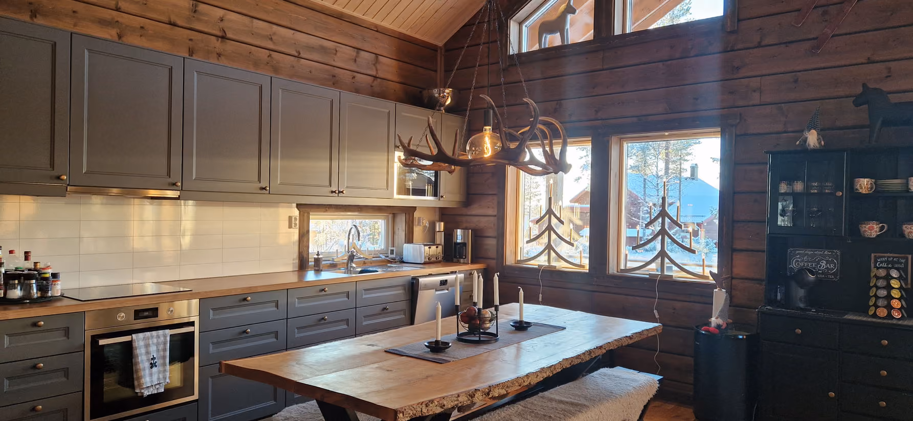 Rustic kitchen with gray cabinets, wooden table, antler chandelier, candle centerpiece, and large windows.