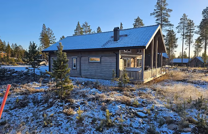 Wooden cabin with a snow-dusted roof and ground surrounded by pine trees under a clear blue sky.