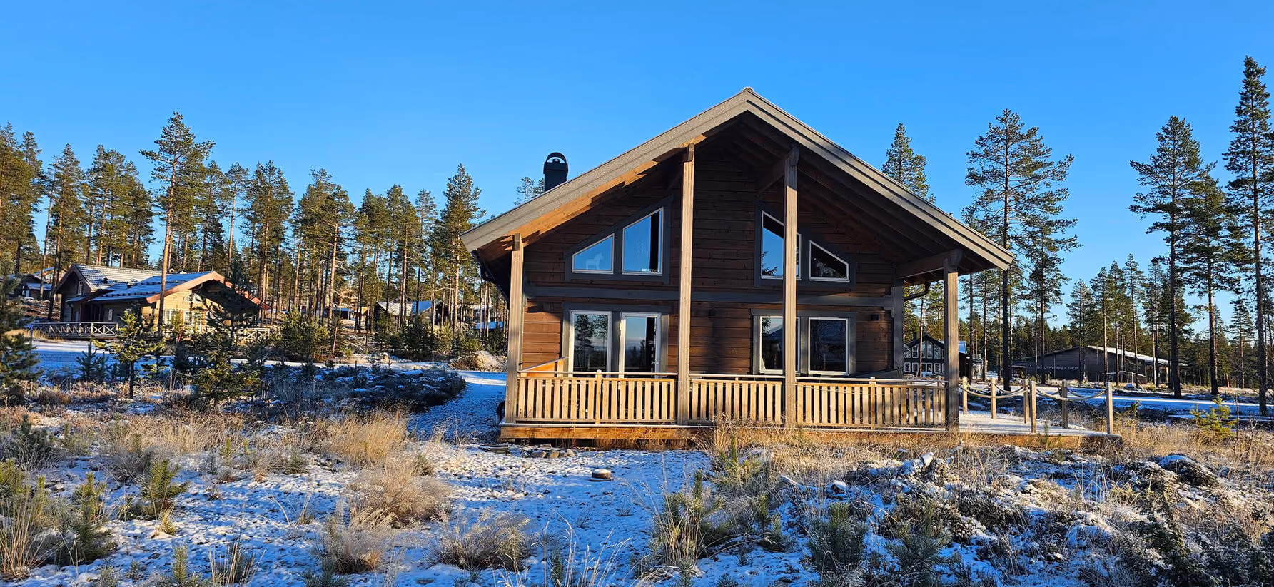 Wooden cabin with porch in a snowy forest clearing under a clear blue sky.
