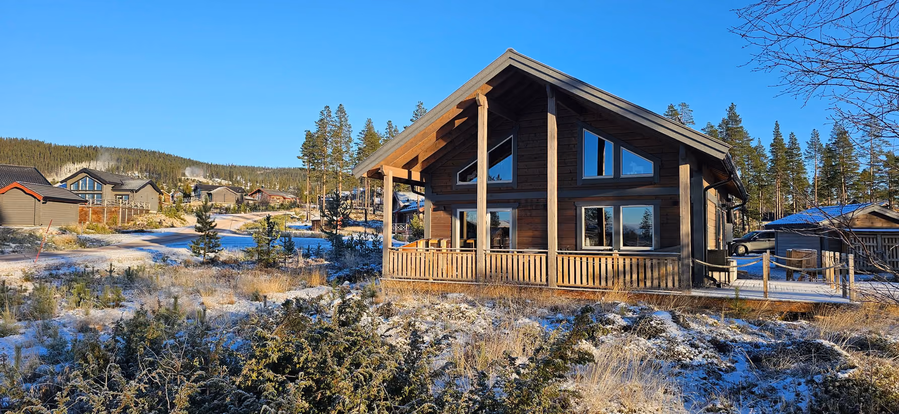 Wooden cabin with large windows in a snowy rural area with pine trees and other houses.