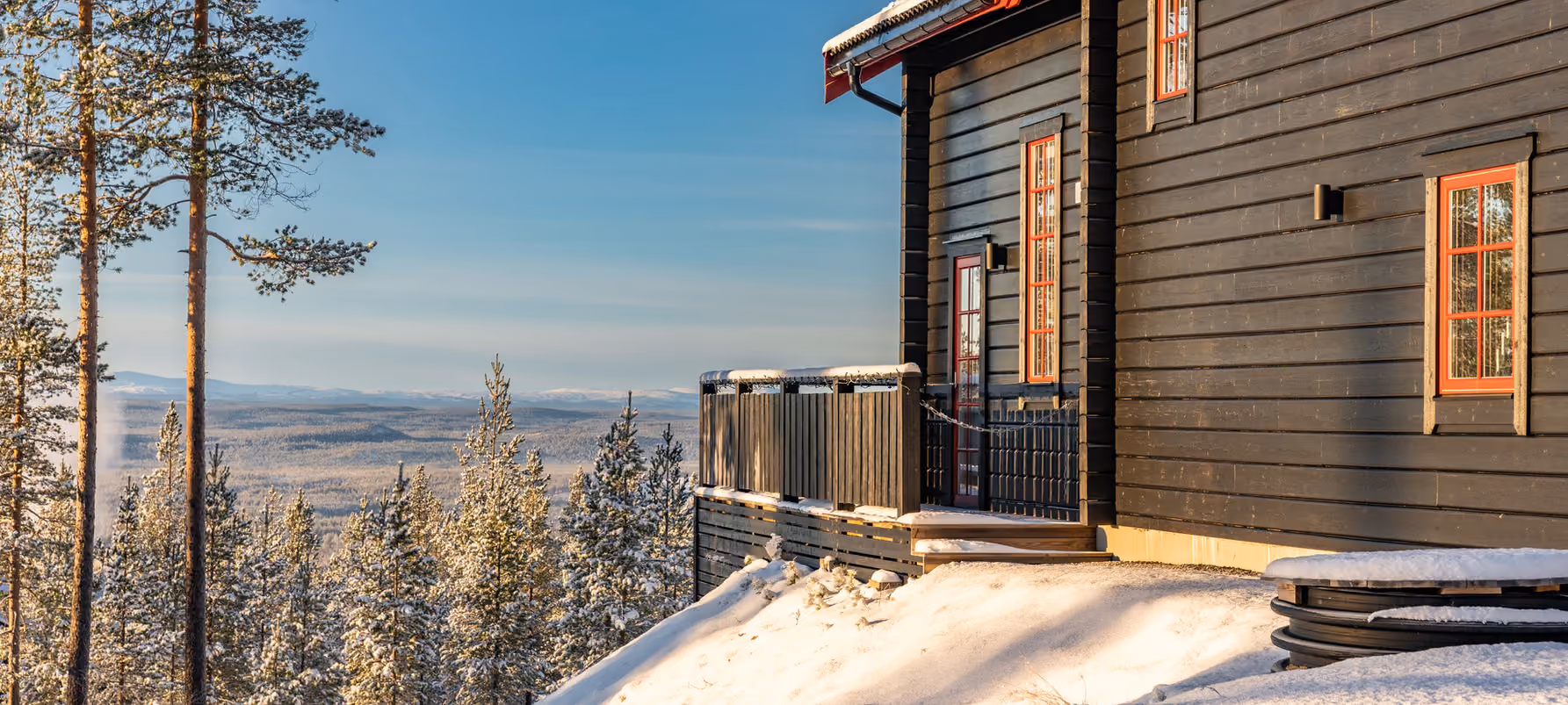 Black wooden cabin with red window frames overlooking a snowy forested valley under a clear sky.