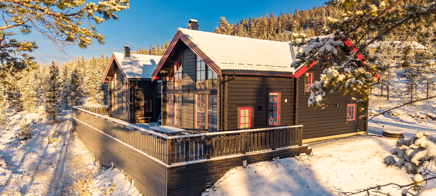 Two black wooden cabins with red window frames covered in snow surrounded by snow-covered pine trees.