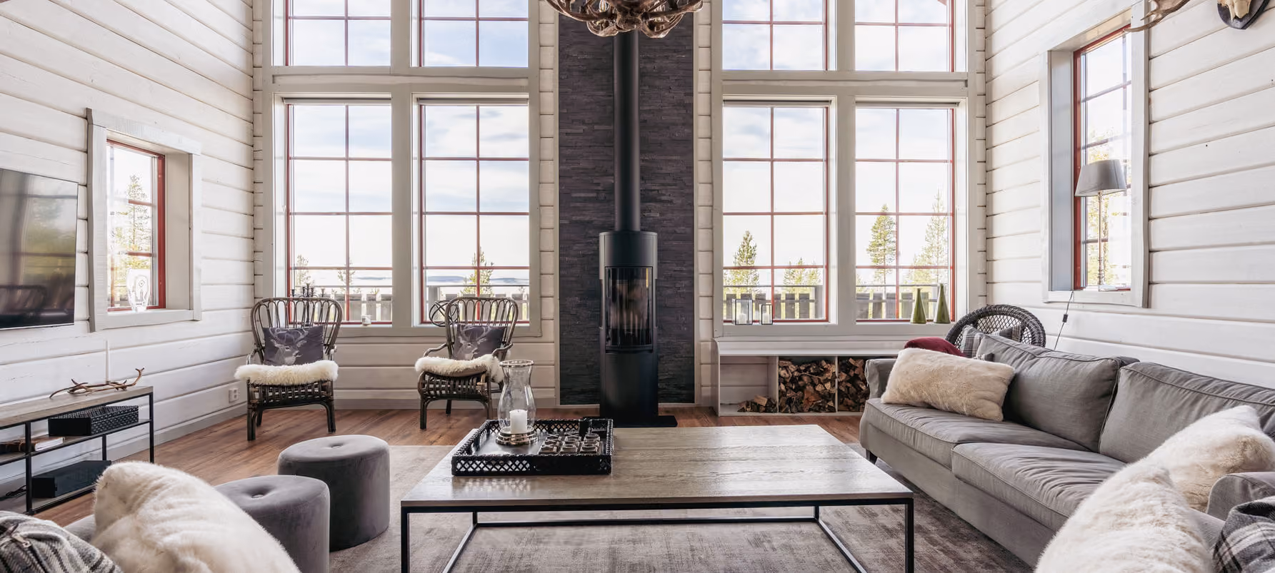 Bright living room with gray sofa, two wicker chairs, a wood stove, and large windows with red frames.