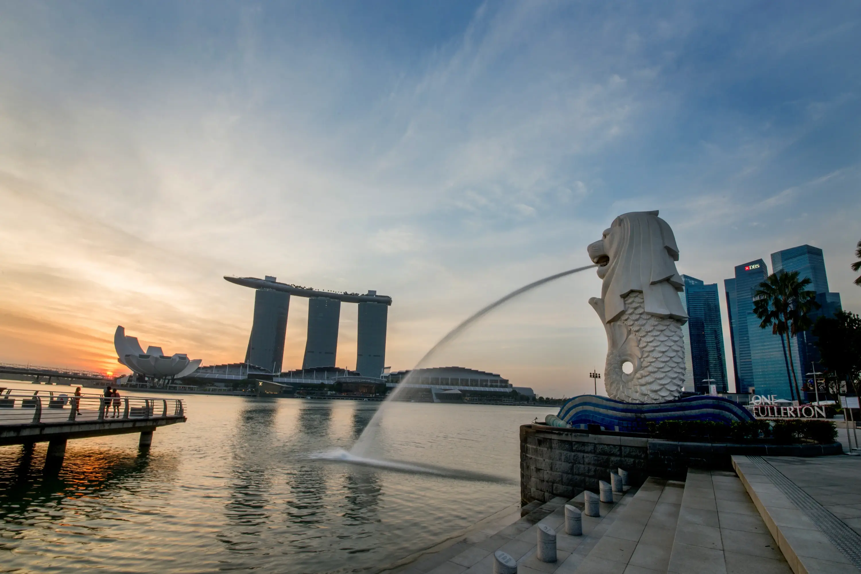 The Merlion statue spouting water into Marina Bay at sunrise, with Marina Bay Sands and the city skyline silhouetted in the distance.