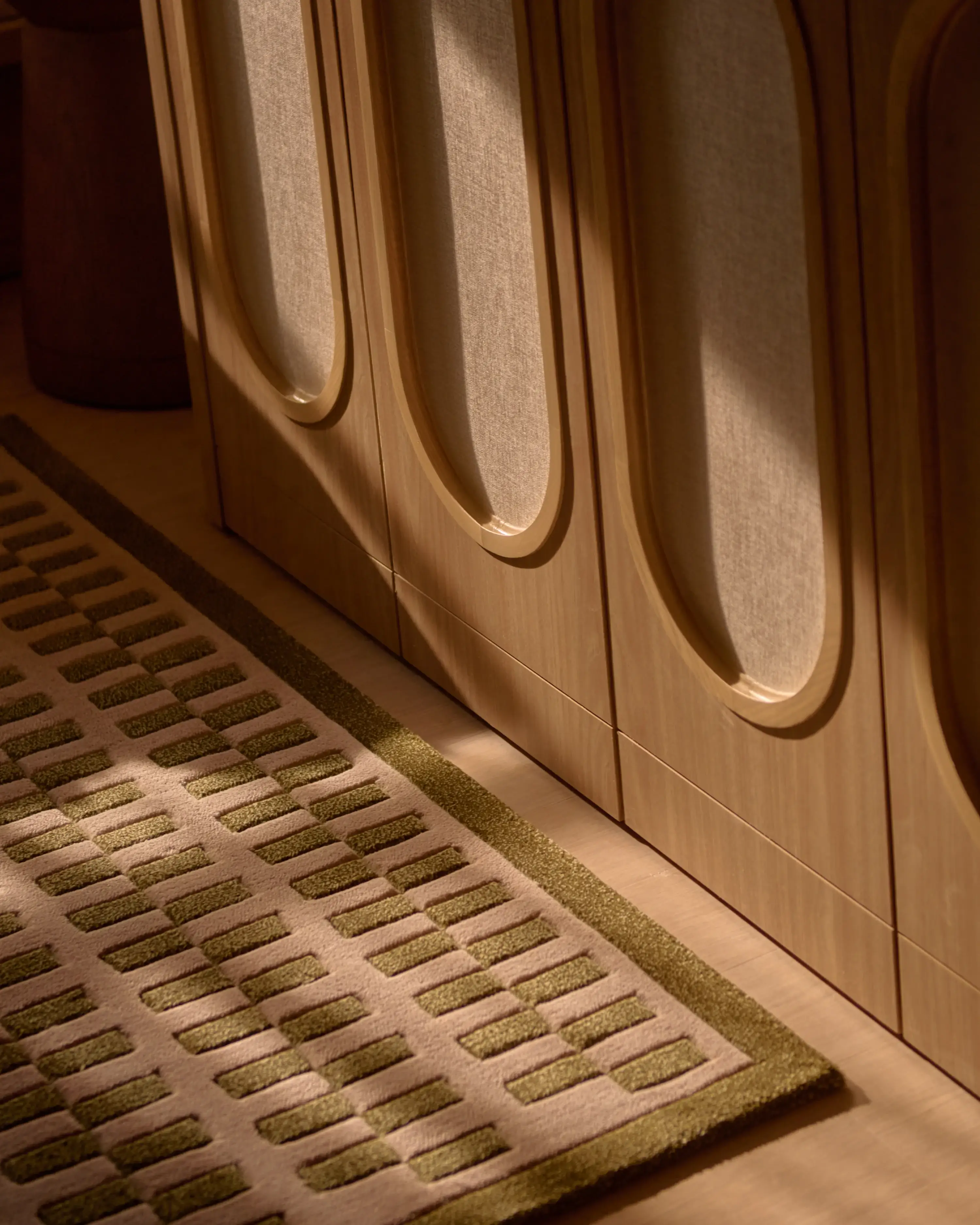 A close-up of a patterned rug in blush and olive tones, laid against the base of a warm wood wardrobe with oval panel detailing, lit by soft afternoon light.