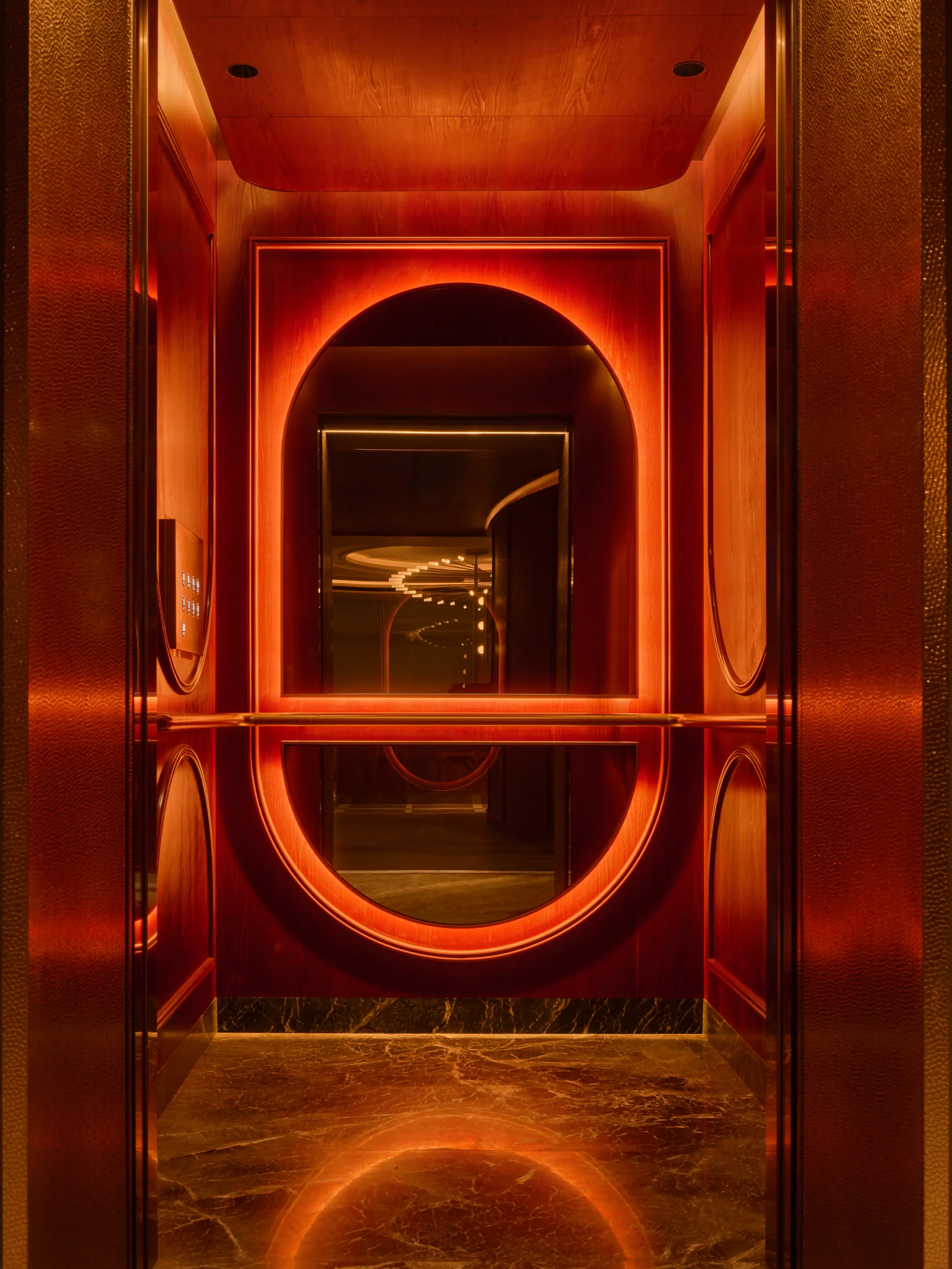 The interior of APT.'s lift, its oval mirror and warm amber lighting creating an infinite reflection against hammered gold walls and marble flooring.