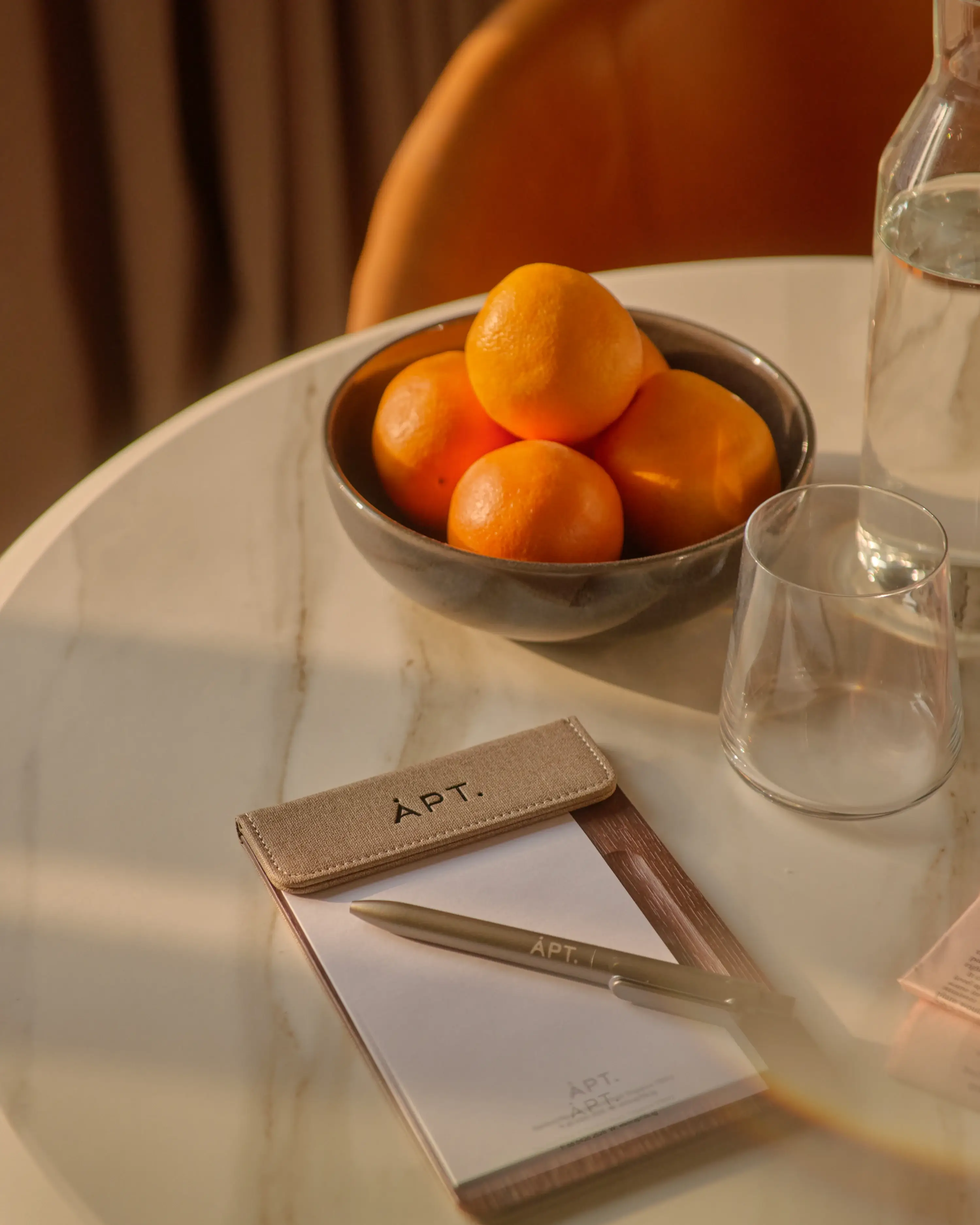An APT. branded notepad, pen case, and a bowl of oranges arranged on a marble dining table, caught in warm afternoon light.