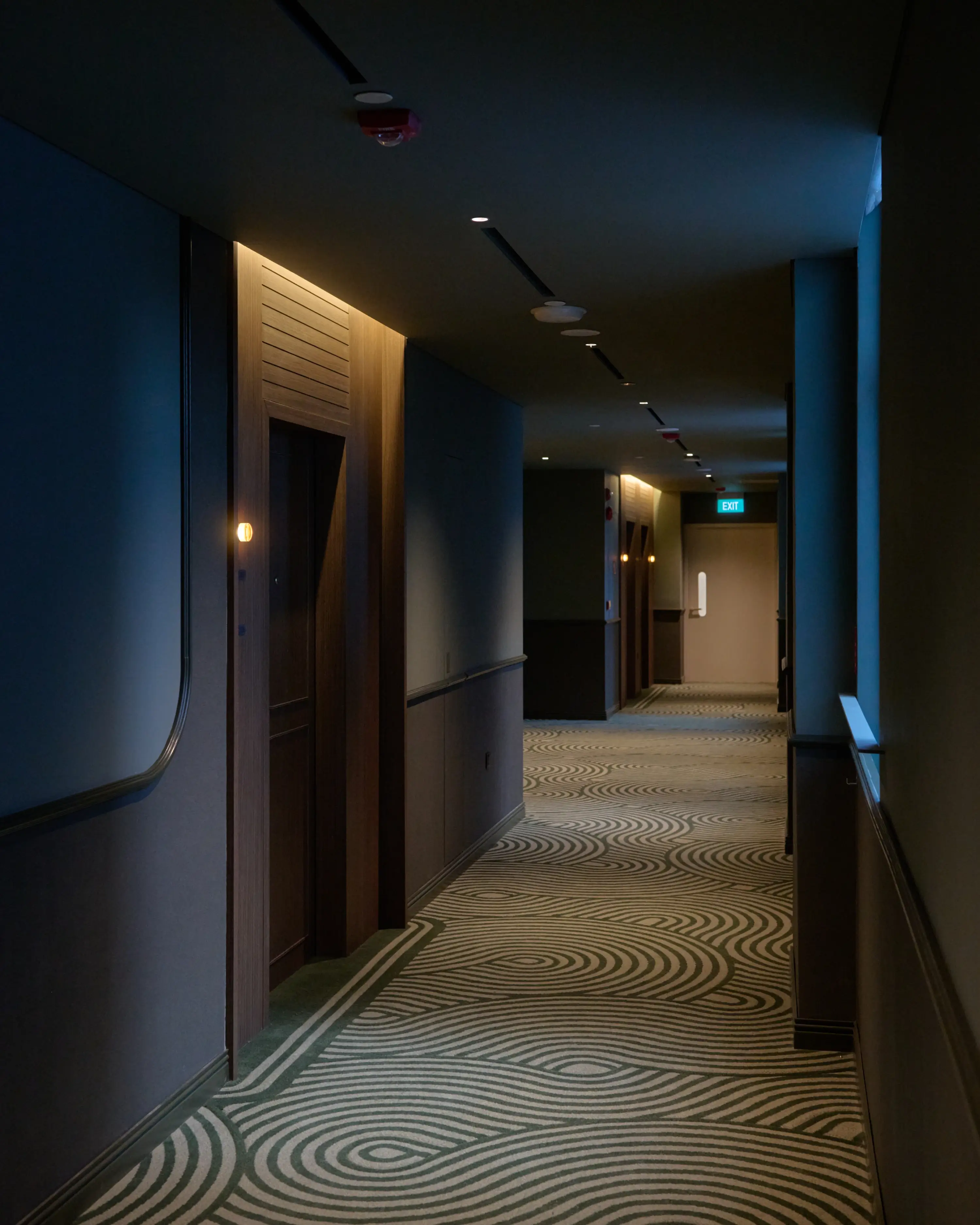 A dimly lit corridor at APT., its swirling patterned carpet stretching into the distance, punctuated by warm amber door lights and dark wood apartment entrances.