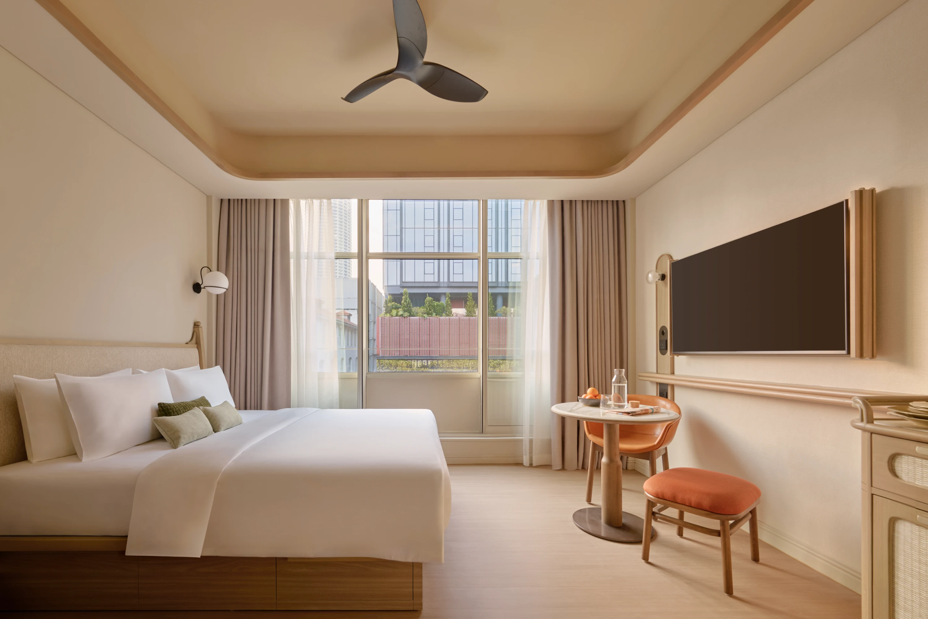 The APT. Studio apartment bedroom bathed in natural light, with a ceiling fan overhead, a round dining table with terracotta chairs, and a city view beyond the window.