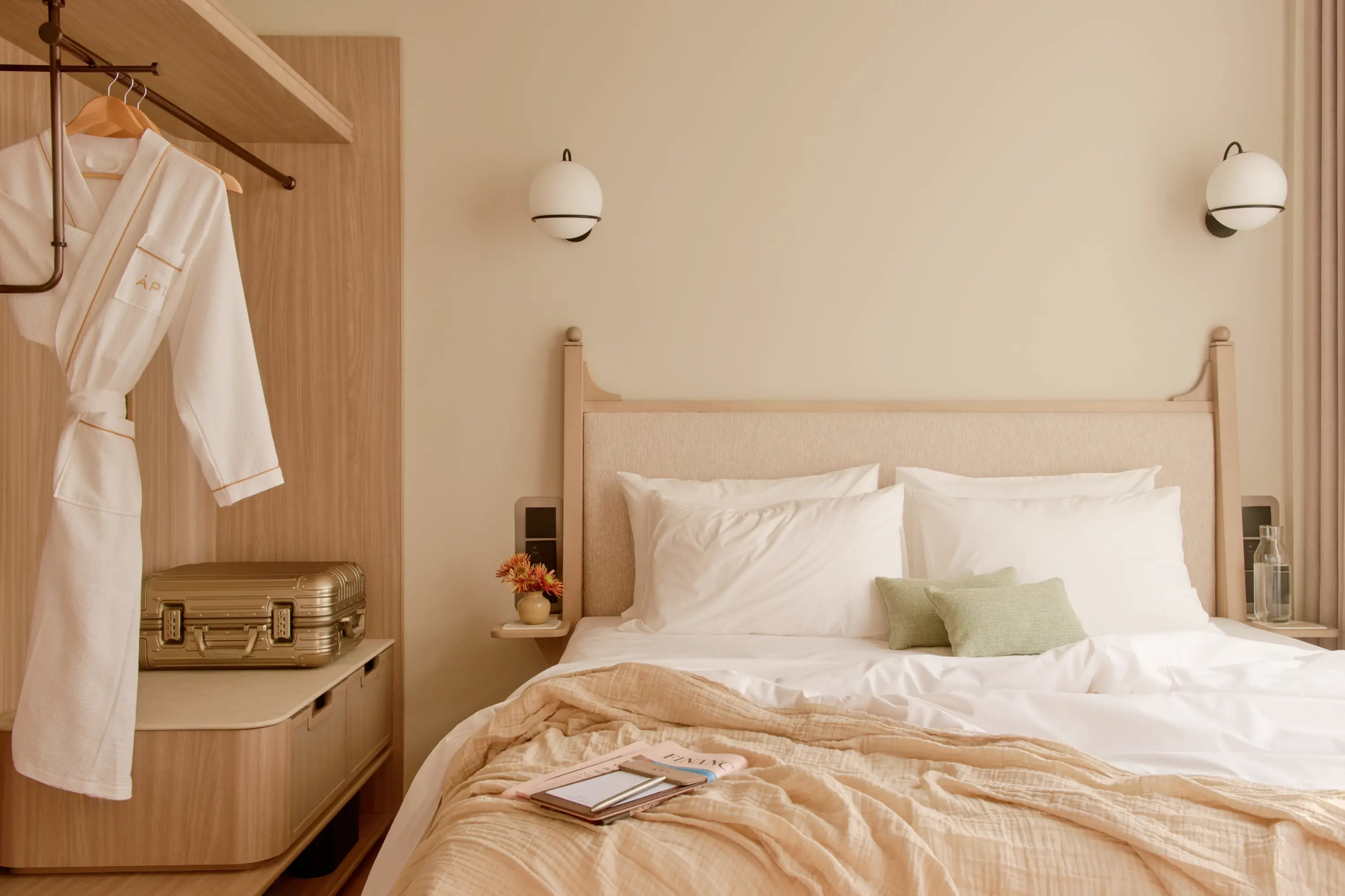 A bathrobe hanging beside an open wardrobe, a gold suitcase resting on the luggage bench, and a neatly made bed with a magazine left open — a lived-in moment in the APT. Studio apartment.