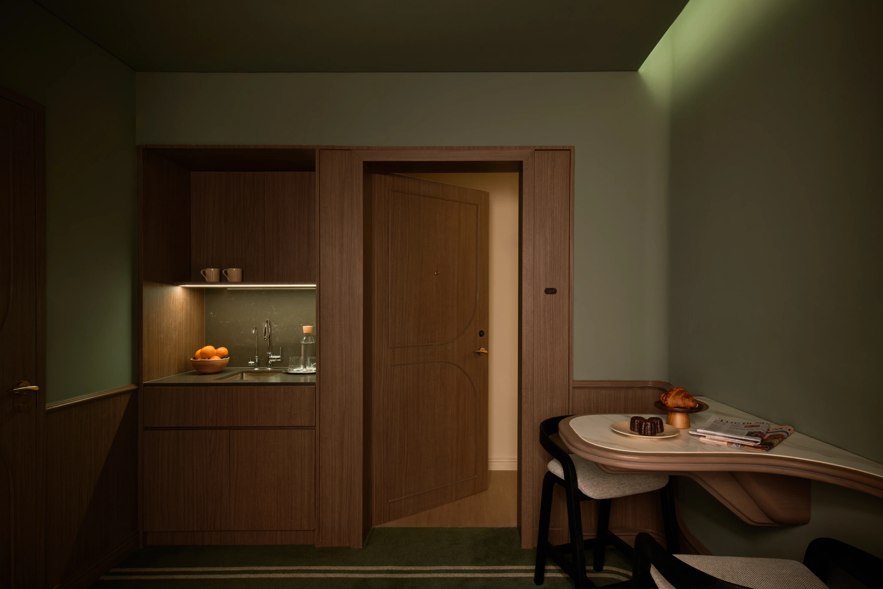 The Vestibule apartment entryway at dusk, its dark wood kitchenette glowing softly under under-cabinet lighting, a croissant and morning reading waiting at the dining nook.