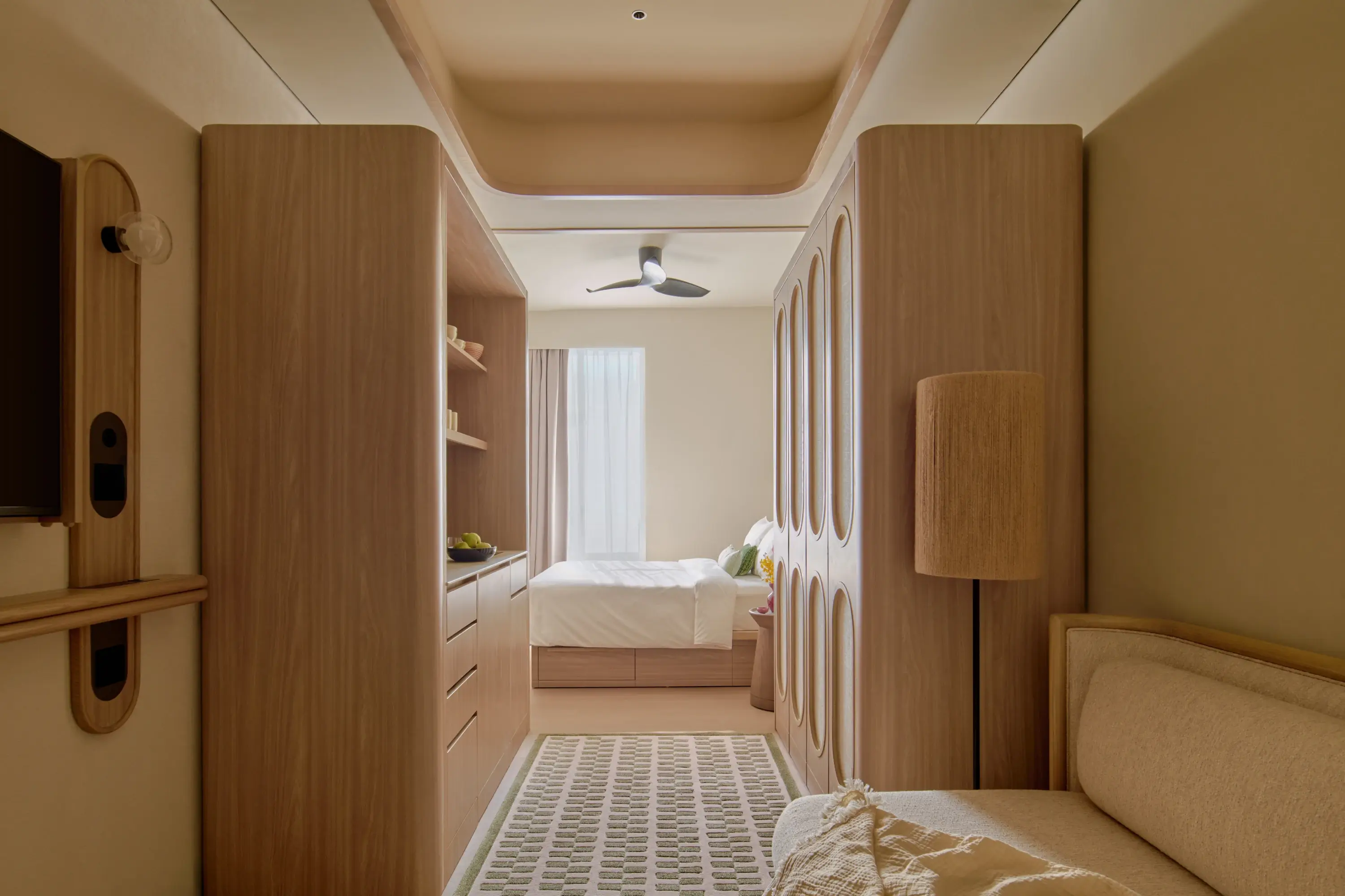 The Deluxe apartment living area framed by warm wood cabinetry, looking through to the bedroom with its oval-panelled wardrobe and ceiling fan beyond.