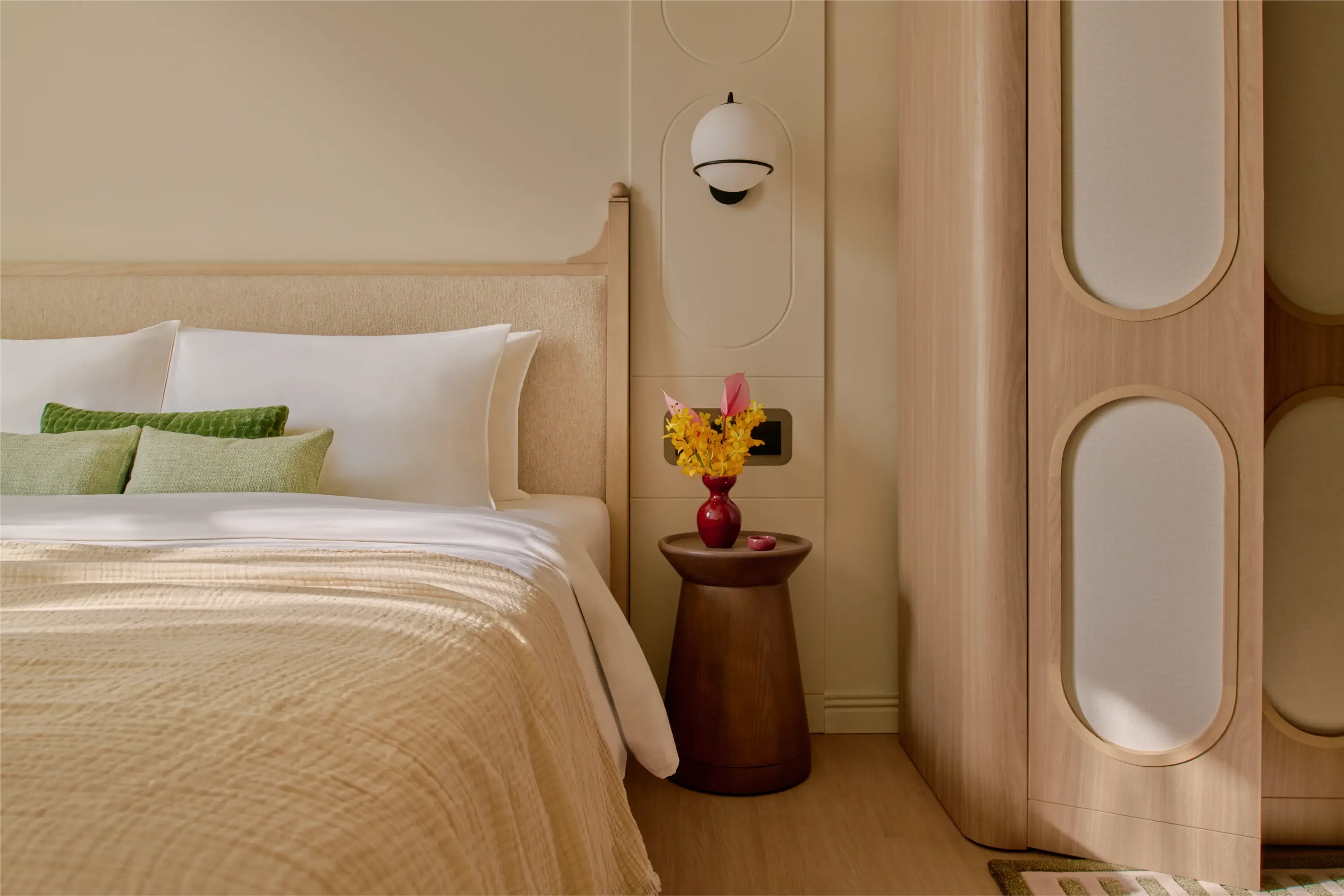 A close-up of the Deluxe apartment bedroom, a red vase of yellow flowers sitting on a dark wood bedside table beside the upholstered headboard and oval wardrobe panels.
