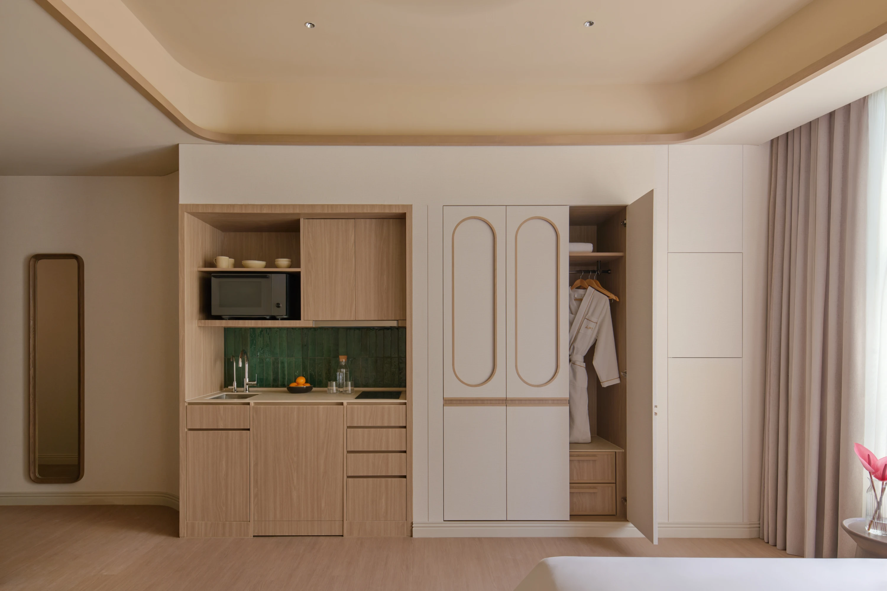 The Urban apartment kitchenette with green tile splashback and microwave, beside an open wardrobe with a bathrobe hanging inside.