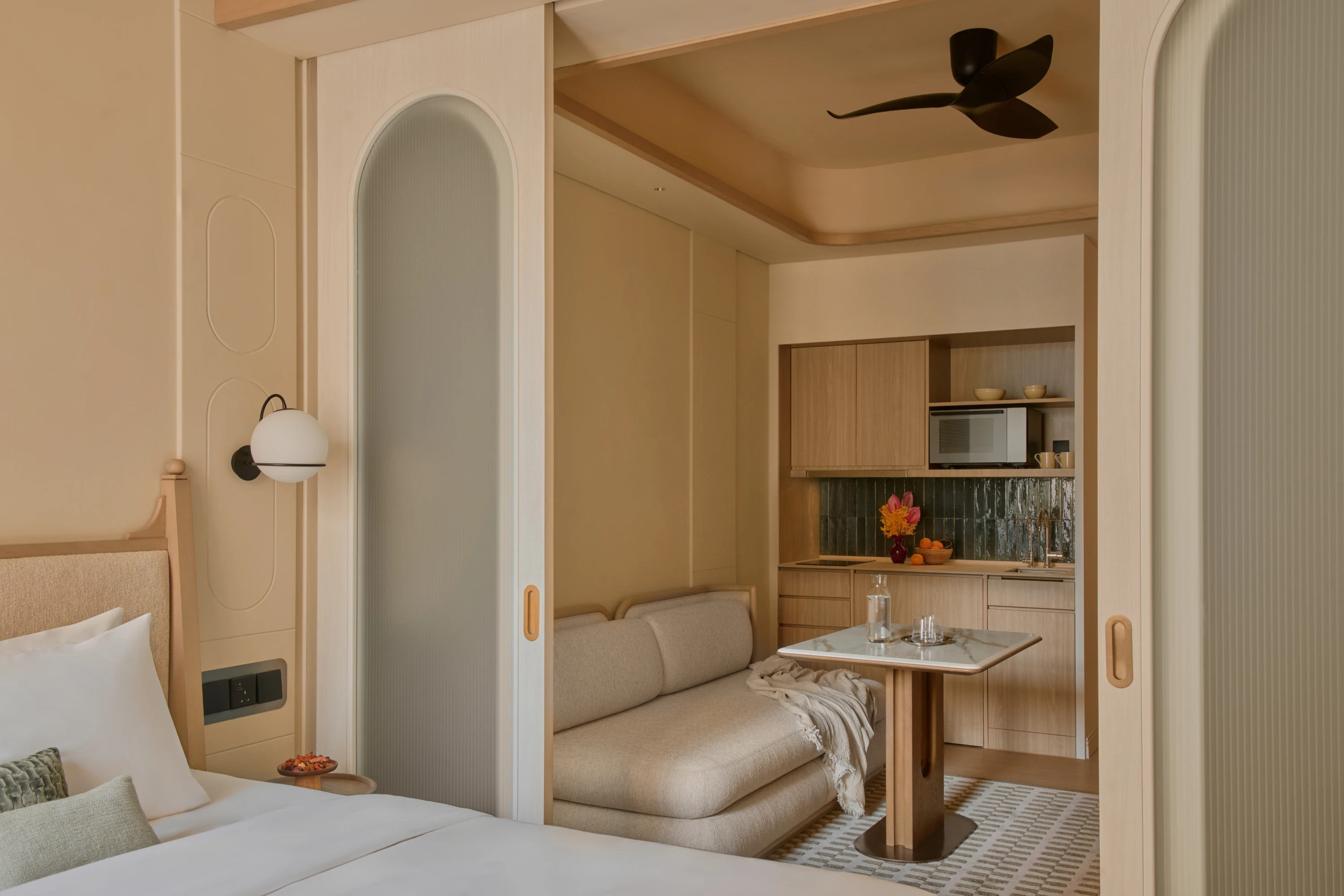 The Stamford Suite bedroom looking through sliding doors into the living area, a sofa and marble dining table set before a kitchenette with green tile splashback.