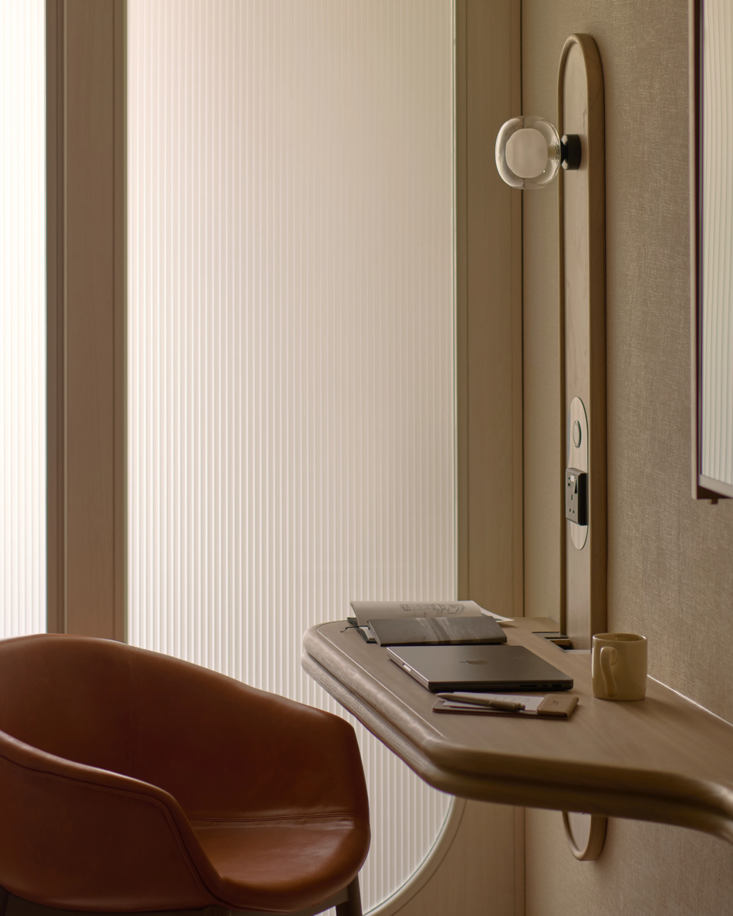 A close-up of the Stamford Premier Suite's wall-mounted desk, a laptop and notepad laid out beside a ceramic mug, lit by soft natural light filtering through ribbed glass panels.