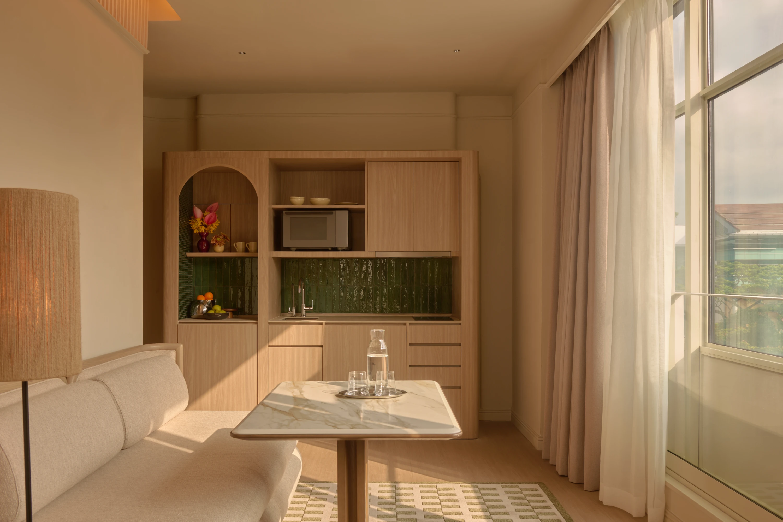 The Civic Suite kitchenette in afternoon sun, light wood cabinetry and a green tile splashback framing an arched display nook with fresh flowers and fruit.