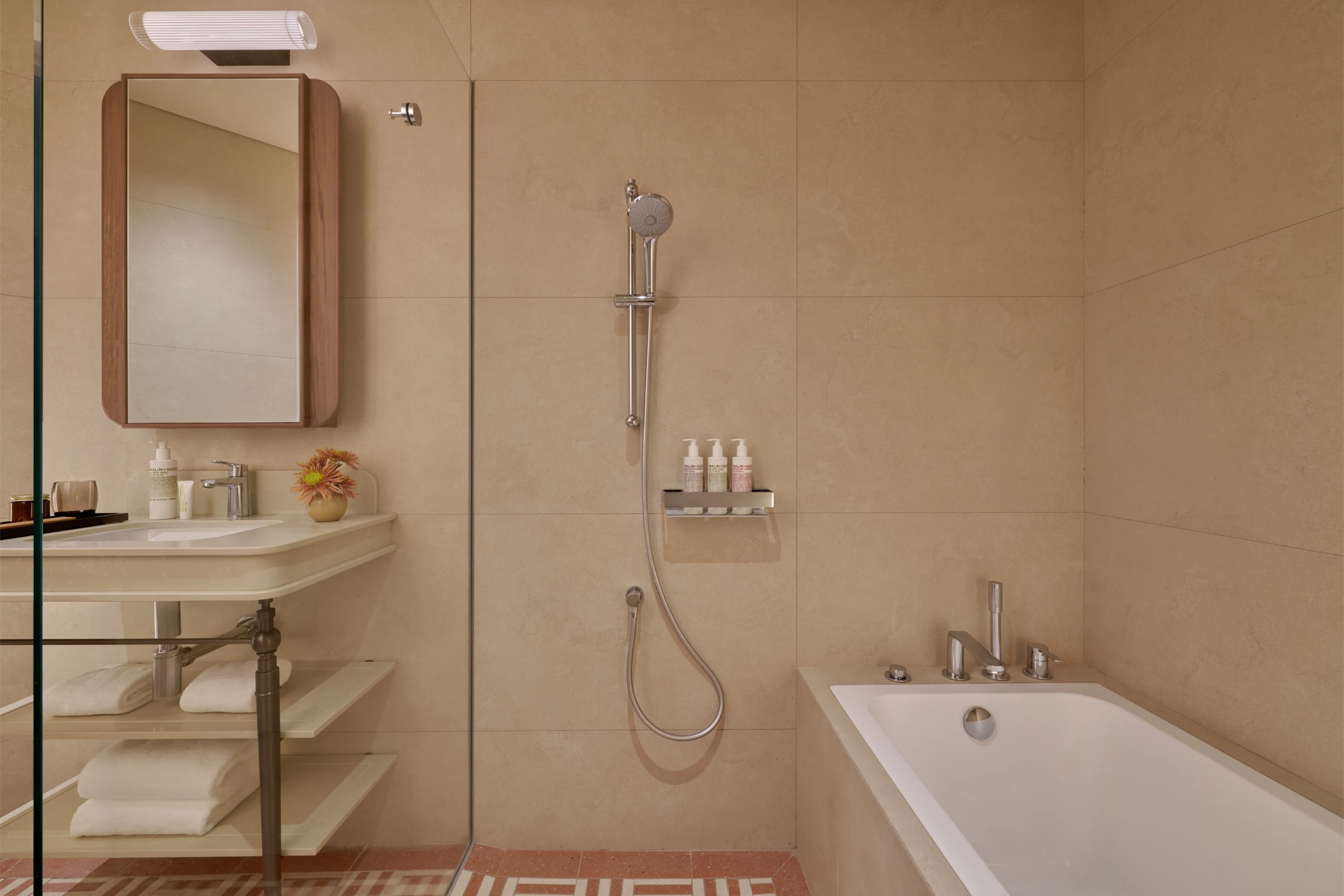 The Civic Suite bathroom with a pedestal vanity, handheld shower, soaking tub, and terrazzo-patterned floor tiles, toiletries neatly arranged on the shower shelf.