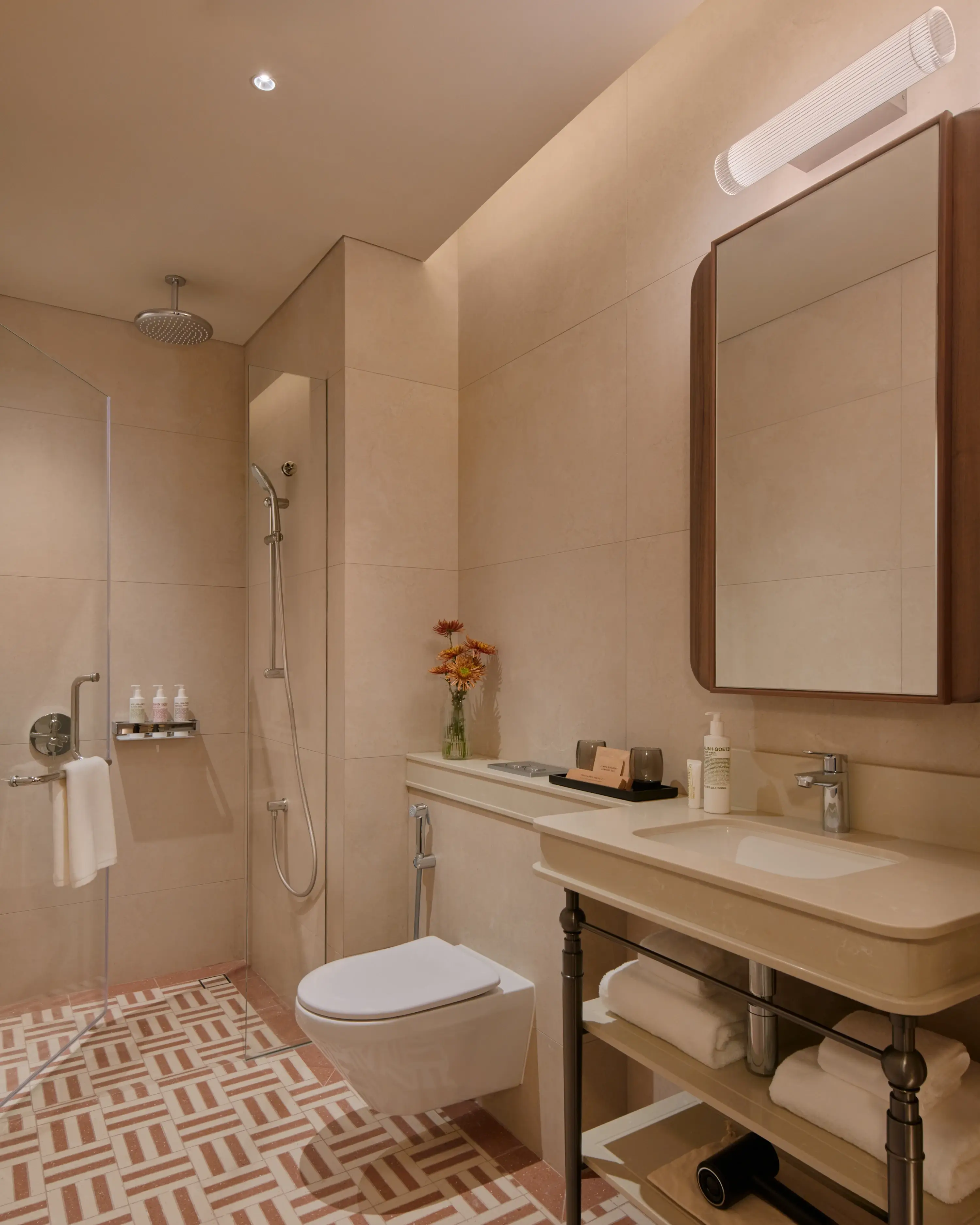 The APT. bathroom with a rain shower, pedestal vanity stocked with toiletries, a small vase of orange flowers on the ledge, and a patterned terracotta tile floor.