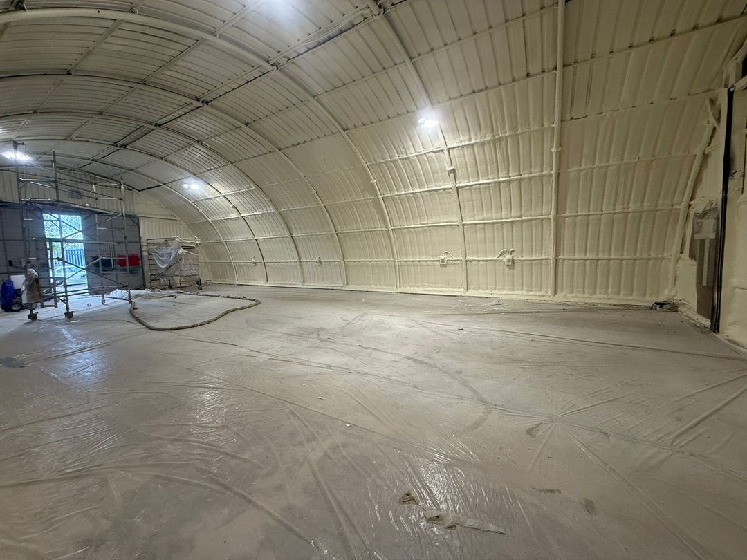 Inside a large arched steel agricultural building: the roof and walls fully sprayed in a continuous cream closed-cell layer, a scaffold tower standing on the masked floor.