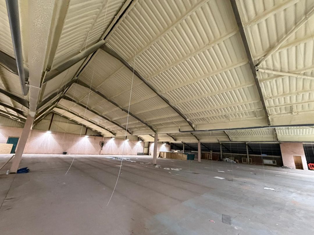 Inside a large single-storey industrial unit: the pitched roof and its steel trusses sprayed from beneath in a continuous cream closed-cell layer, the concrete floor cleared beneath it.