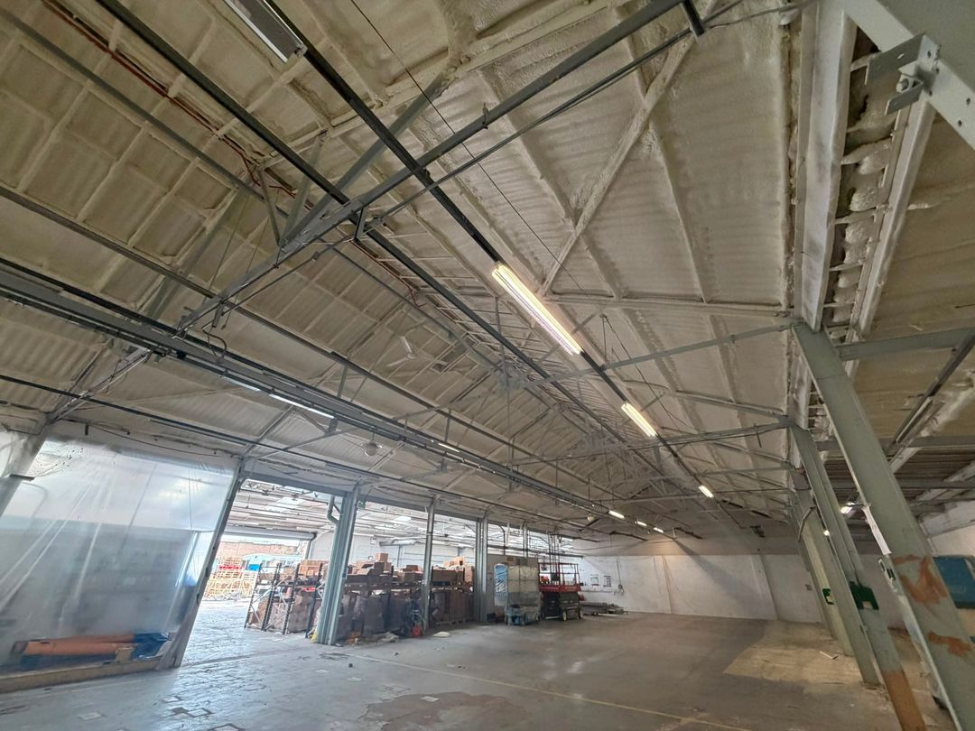 A large warehouse truss roof sprayed from beneath in closed-cell foam, polythene screening and stock at the far end.