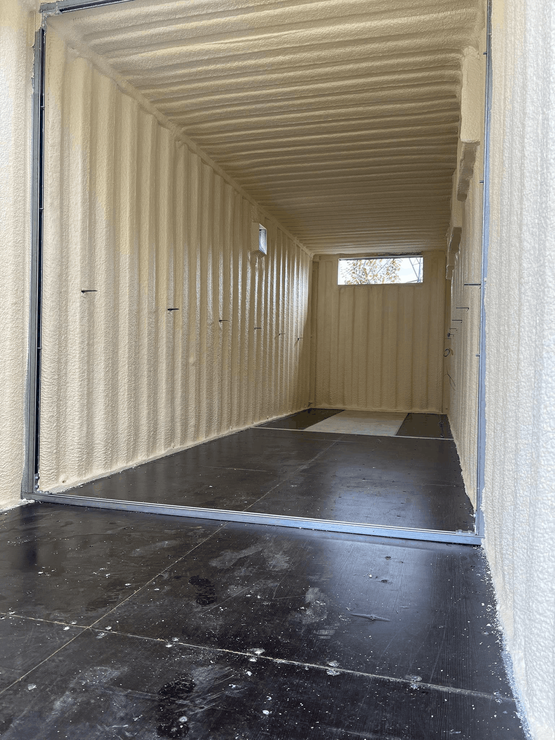 A shipping container conversion interior sprayed with closed-cell foam, walls and roof in one continuous cream layer.