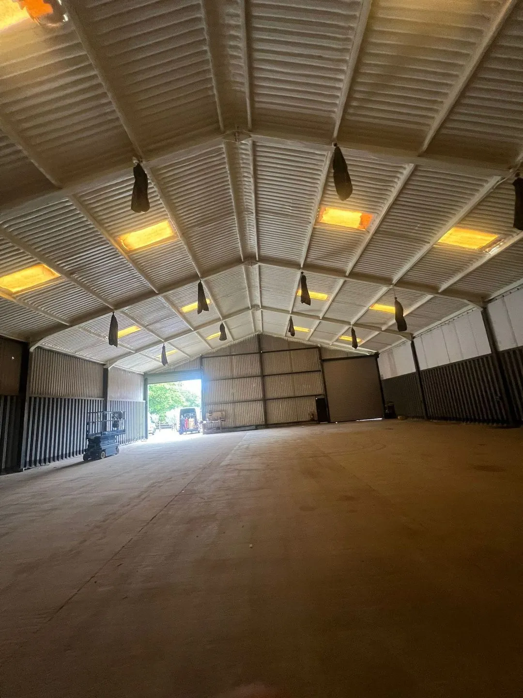An empty warehouse floor looking towards an open roller-shutter door: the whole profiled steel roof and its purlins sprayed from beneath in a continuous pale closed-cell layer, rooflights and high-bay lights left exposed.