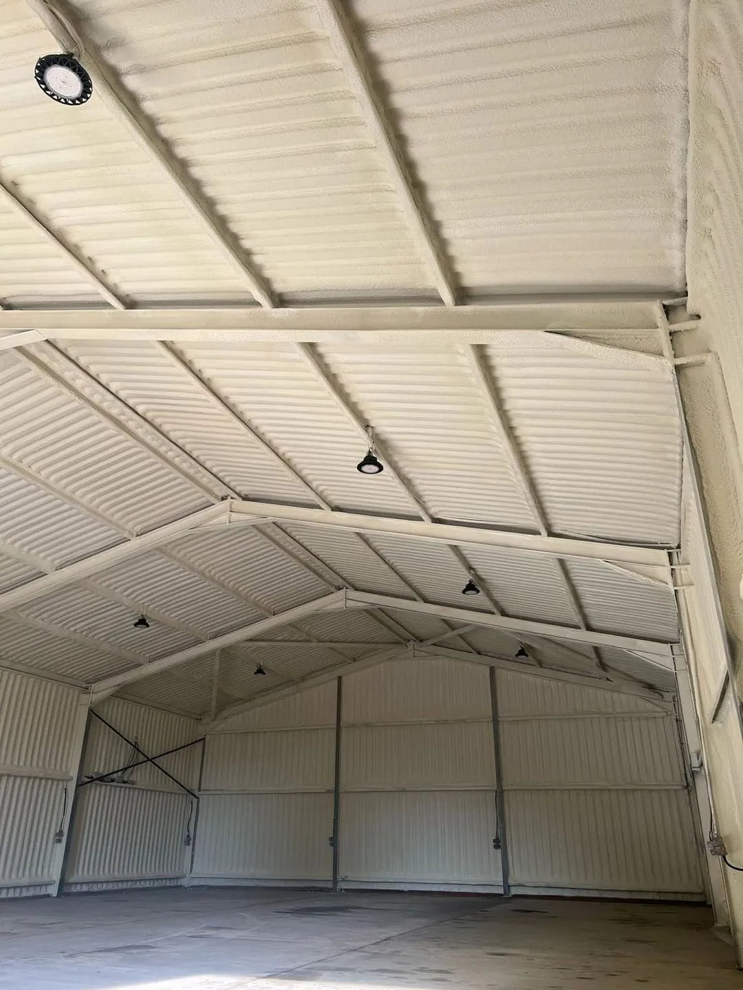 A corrugated metal roof sprayed from beneath in a continuous pale closed-cell layer, seen from inside the building.