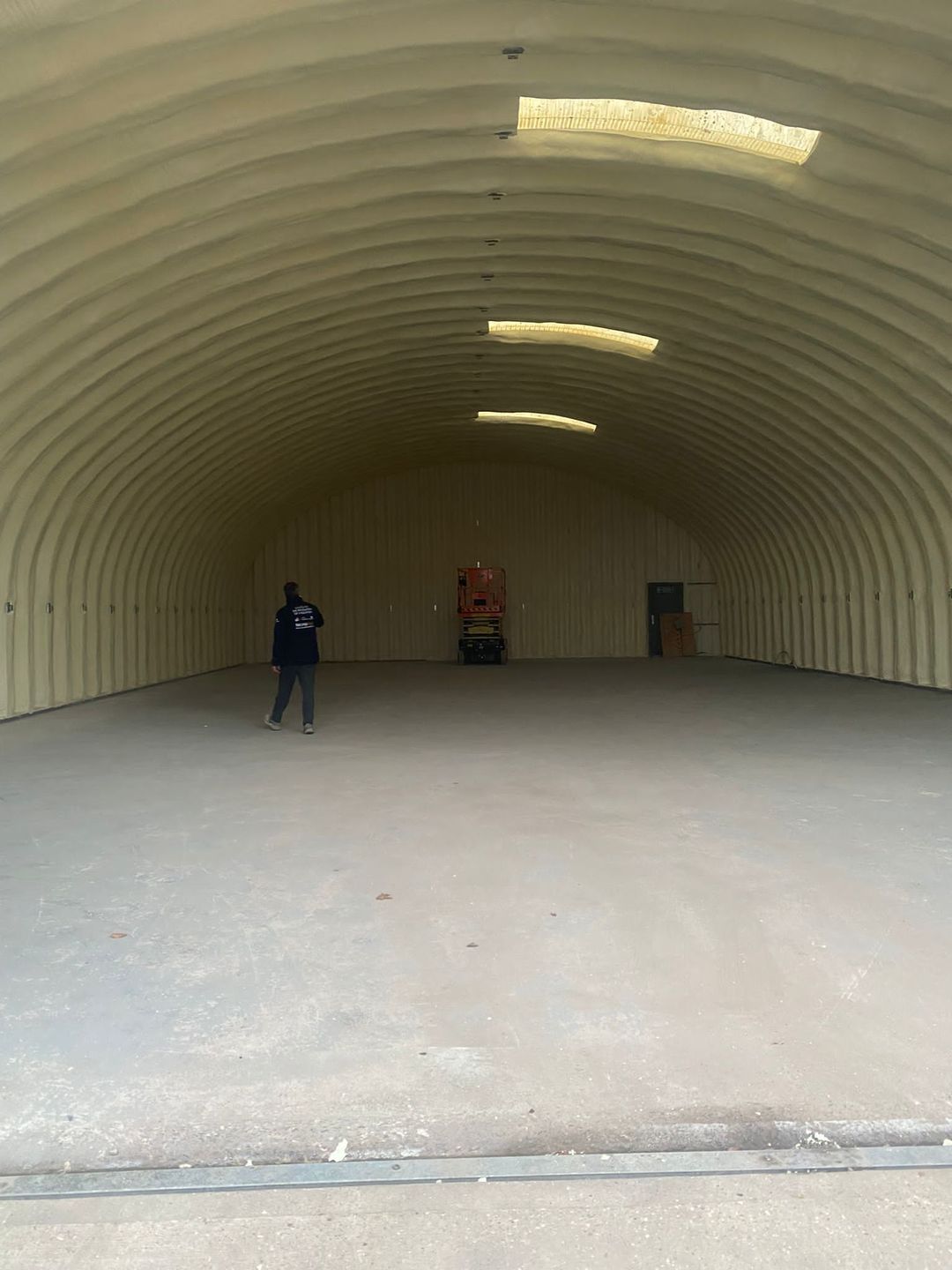 Inside an arched Nissen hut: the whole curved underside sprayed in a continuous cream closed-cell layer, rooflights left clear, a scissor lift at the far end.