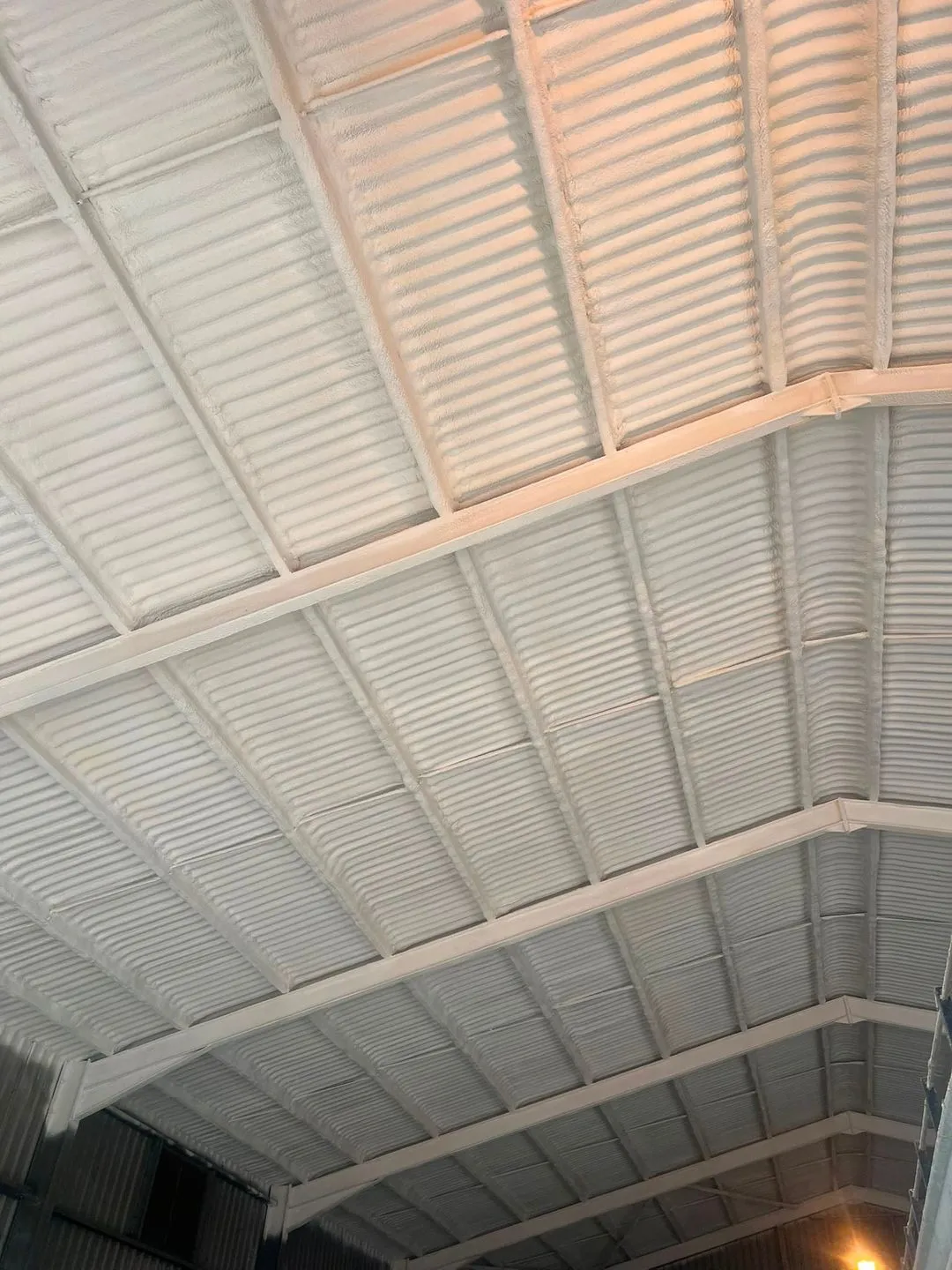 A metal roof sealed from beneath with a continuous pale closed-cell layer, seen from the floor of the building.