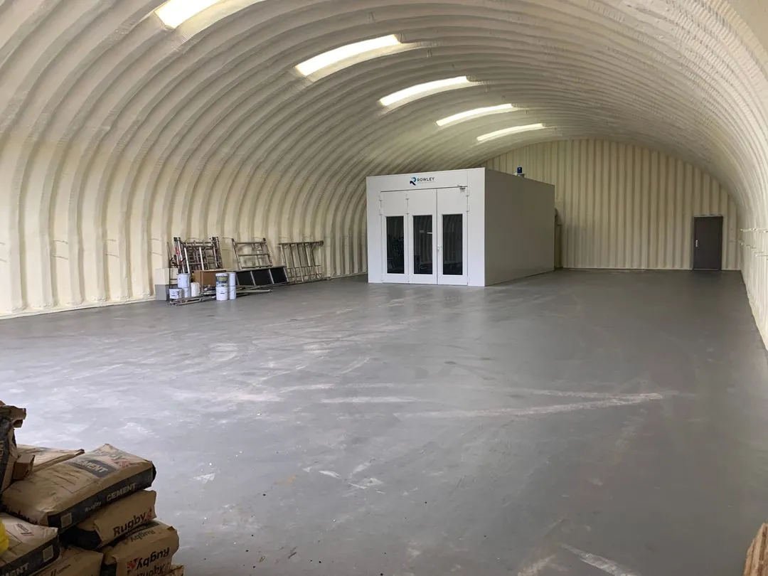 Inside a finished arched steel agricultural building: the curved roof and walls sprayed in one continuous cream closed-cell layer, a spray booth installed inside, bags stacked on the grey floor.