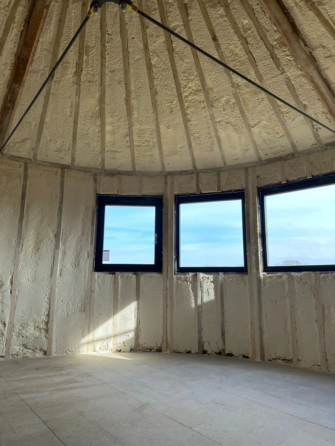 Inside a round silo: the walls and the conical roof sprayed in a continuous cream closed-cell layer between timber studs, three windows letting in daylight.