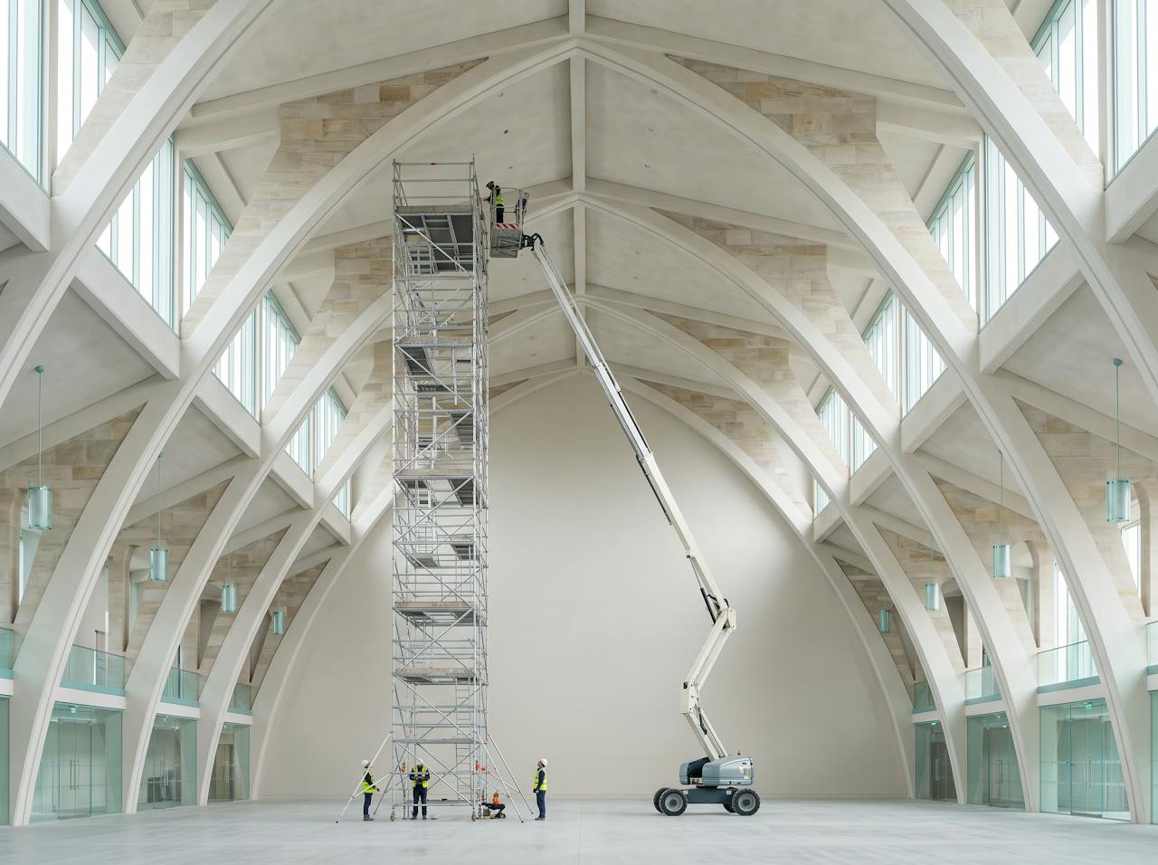 A tall arched hangar roof seen from the floor, scaffold towers and a mobile elevating work platform reaching up to the curved underside, pale daylight through the end doors.