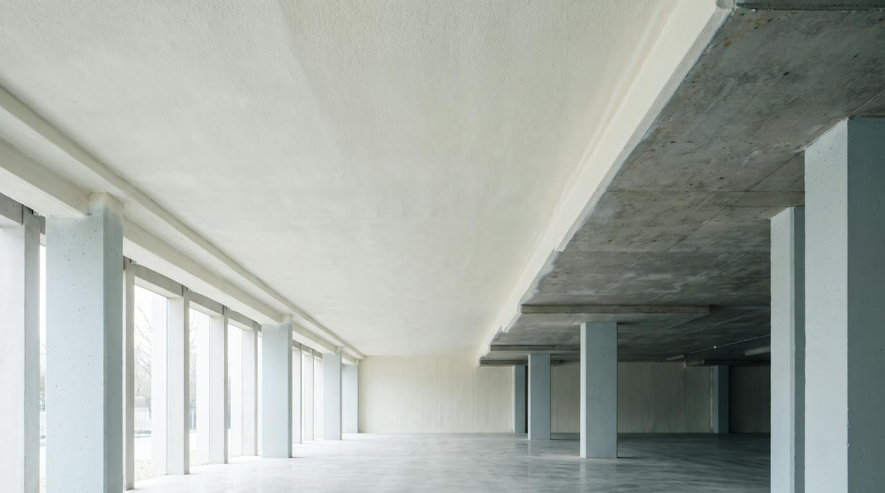 The underside of a concrete floor slab over an open undercroft car park: bare grey soffit, downstand beams, a row of columns and pale daylight from the open side.