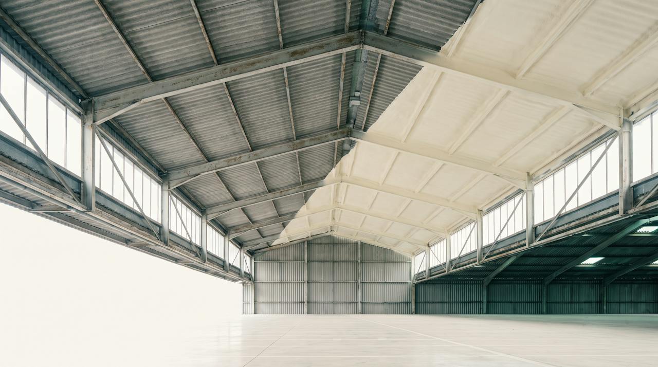 The underside of a large profiled industrial roof seen from the floor, one half sealed in a continuous pale closed-cell layer and the other still bare sheet, pale daylight along the purlins.