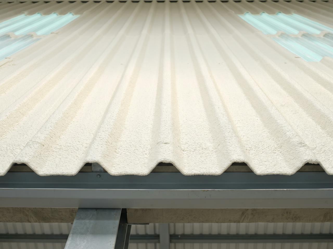 The underside of a corrugated roof with a continuous cream closed-cell layer following every corrugation, the purlins wrapped, raking daylight showing the even finish.
