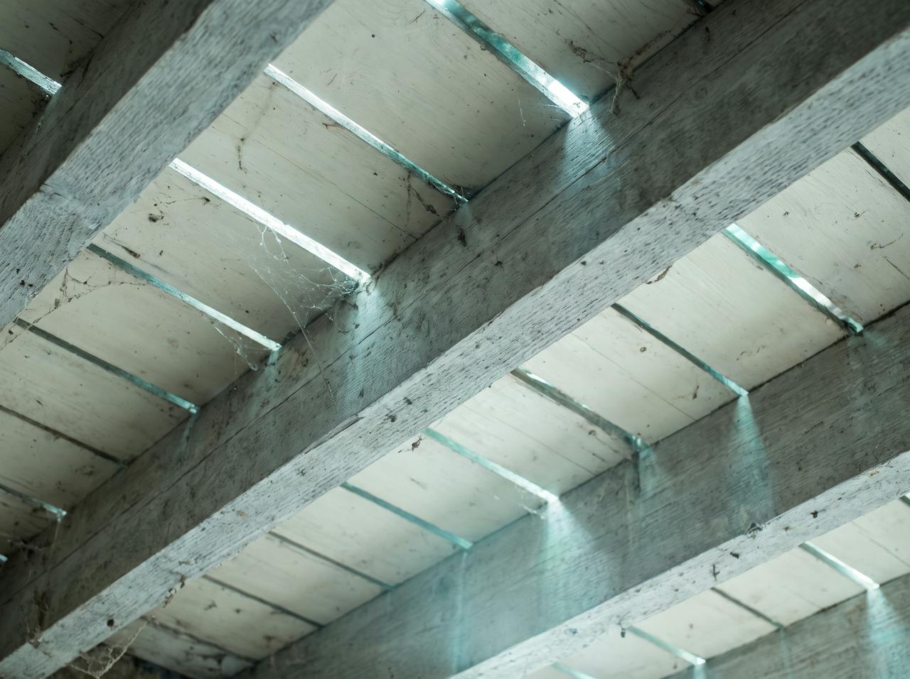 A close detail of the underside of bare floorboards between two joists, seen from the void, gaps of light at the board joints and cobwebs on the timber.