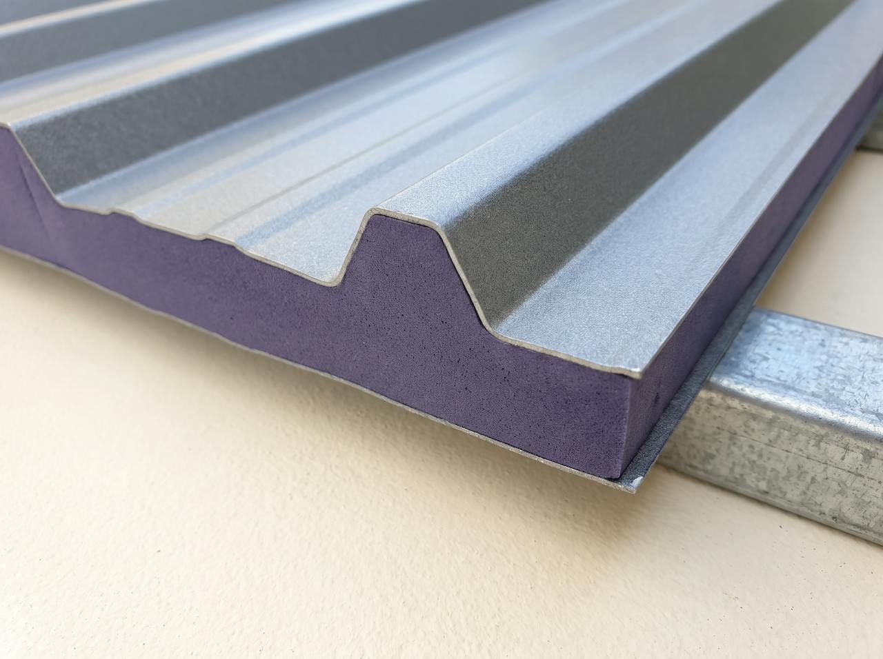 A cut sample of profiled steel roof sheet on a purlin, with a closed-cell layer applied to its underside following the profile, cut cleanly to show the section.